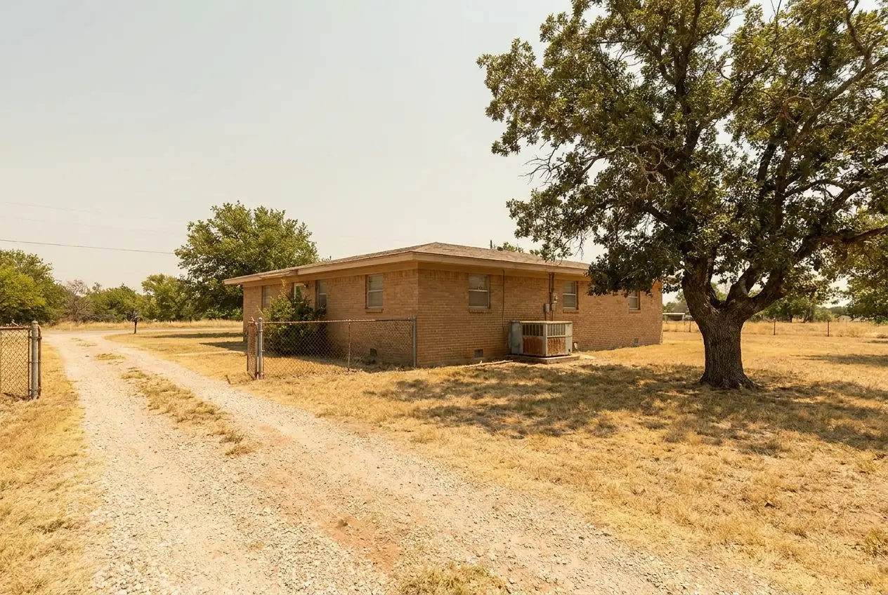 A brick ranch house on a rural lot, an air conditioning condenser at its side and an oak in the yard
